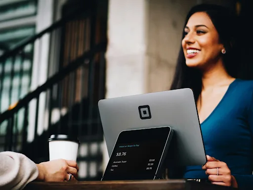 Smiling cashier using a tablet-based payment system to assist a customer holding a coffee cup.
