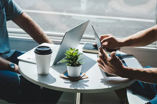  Two people working on laptops at a small café table with a coffee cup and plant.
